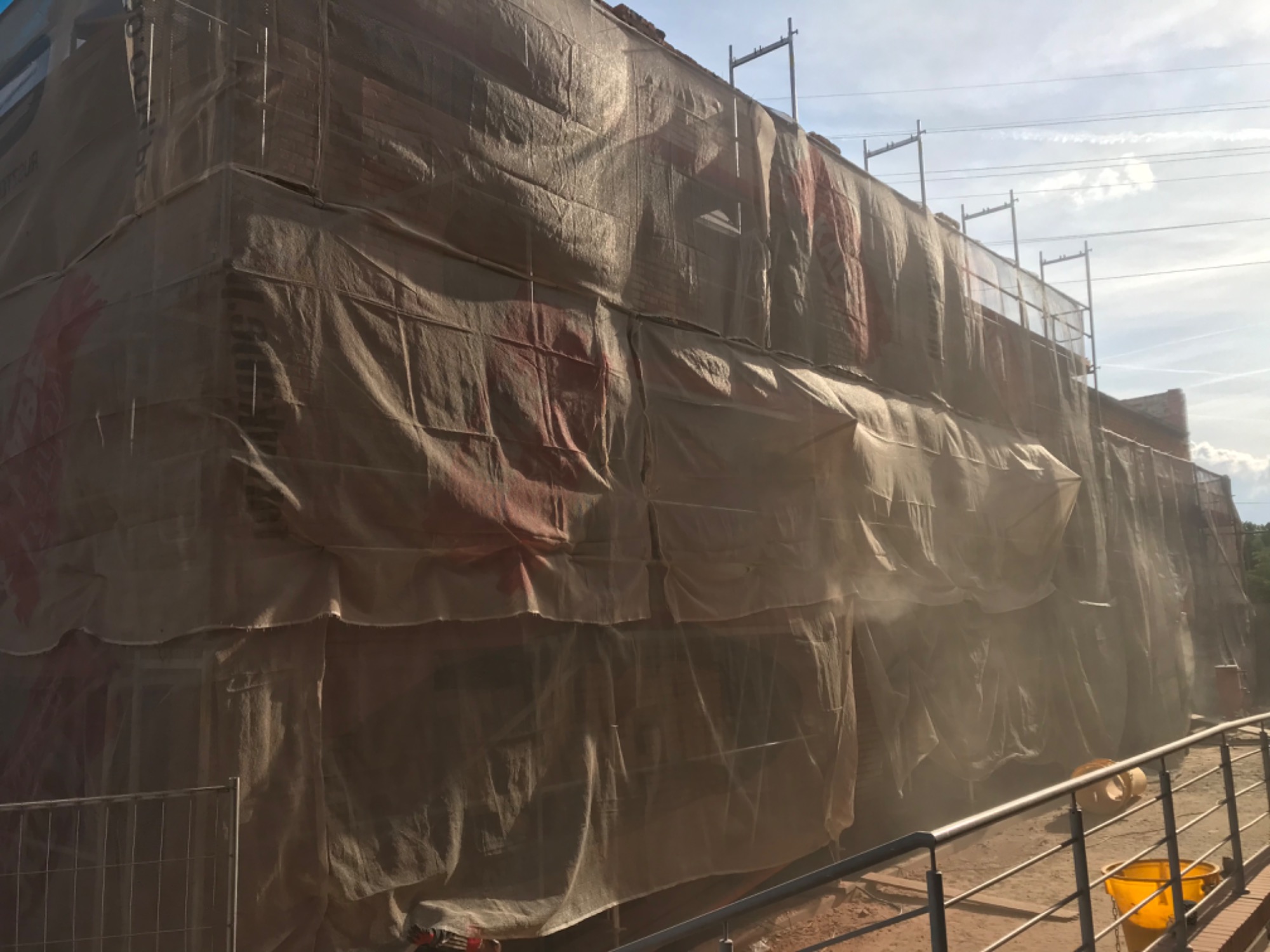 Budynek w trakcie prac elewacyjnych, osłonięty siatką ochronną na rusztowaniu, widoczny fragment chodnika z żółtym wiadrem.