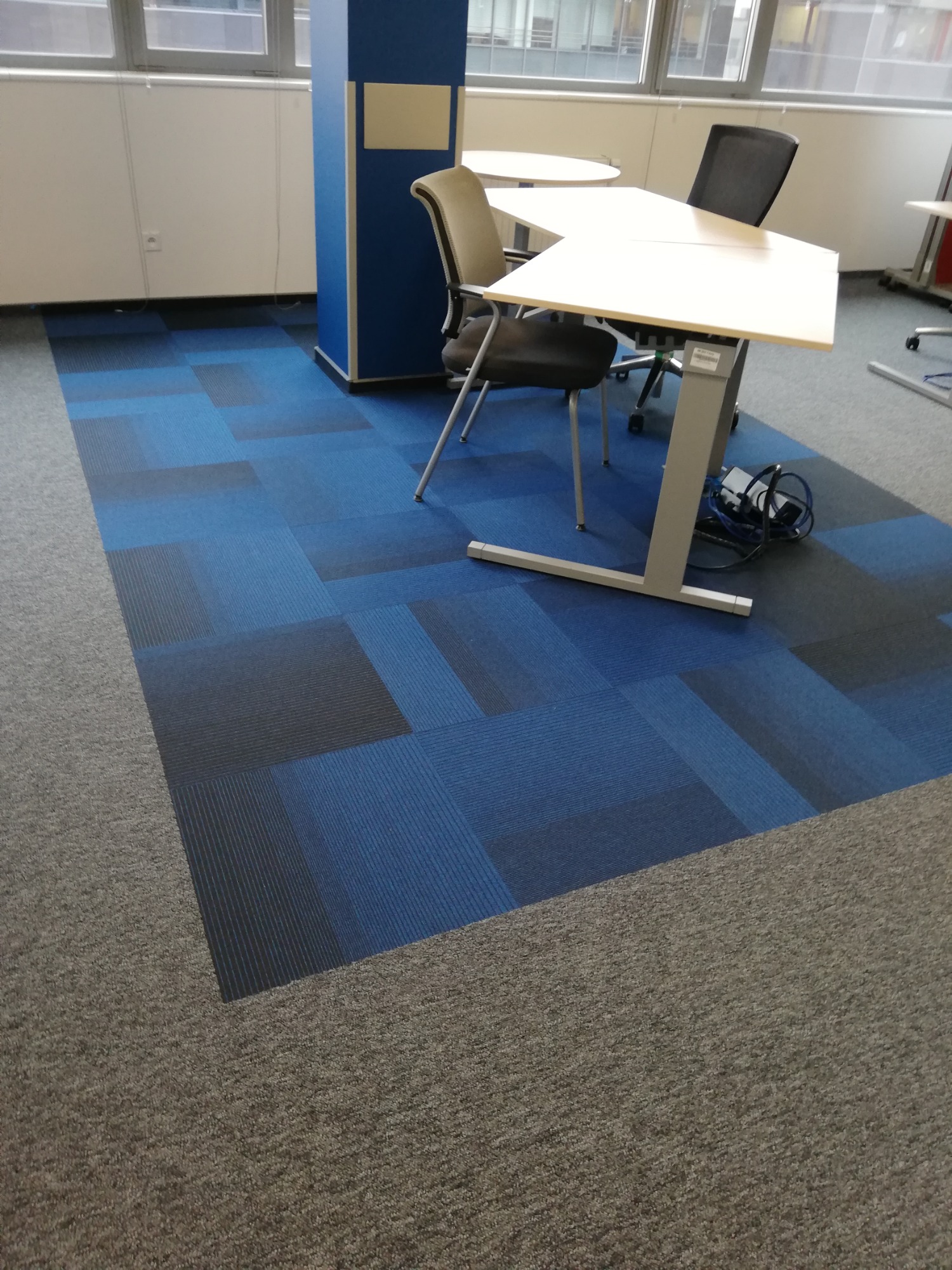Biuro z podłogą wyłożoną kwadratowymi panelami wykładzinowymi w odcieniach szarości i błękitu, widoczne biurka i krzesła.