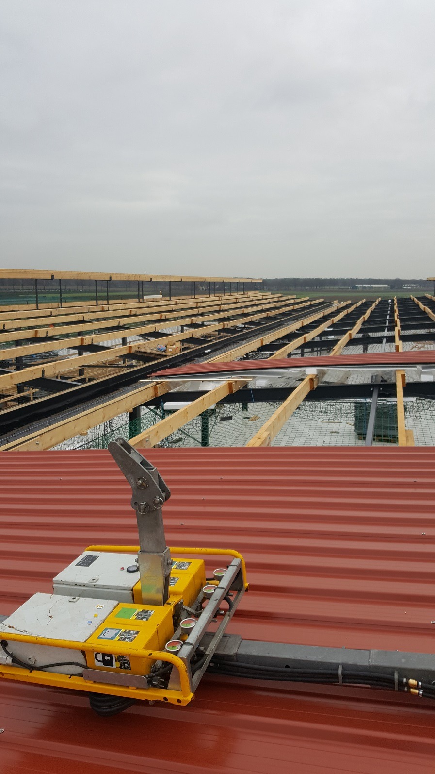 Czerwony dach z blachy trapezowej z widocznym urządzeniem do podnoszenia płyt, w tle konstrukcja drewniana hali w trakcie budowy, pochmurne niebo.
