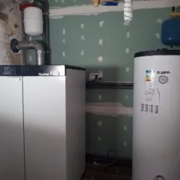 Nowoczesny piec gazowy Daspell Nex i bojler Kospel z niebieskim zbiornikiem wyrównawczym w surowym pomieszczeniu z częściowo wykończonymi ścianami.