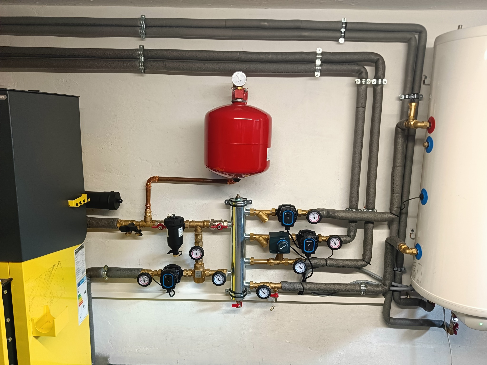 Kotłownia z widocznym kotłem, czerwonym zbiornikiem wyrównawczym, białym zasobnikiem ciepłej wody oraz rozbudowaną instalacją hydrauliczną z licznymi zaworami, pompami obiegowymi i manometrami.