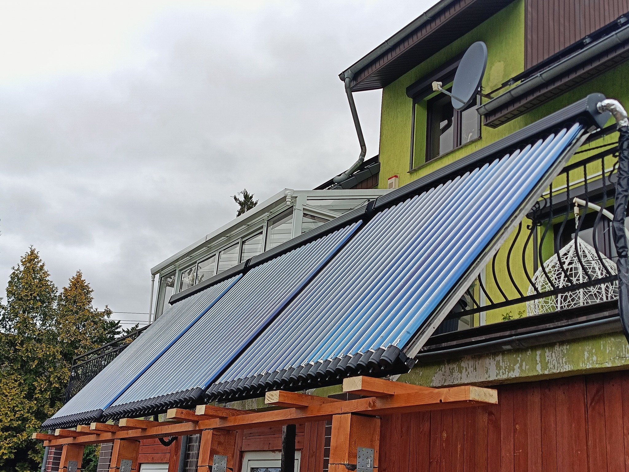 Kolektory słoneczne zamontowane na drewnianej konstrukcji przy zielonym domu z satelitą, widok z dołu w pochmurny dzień.
