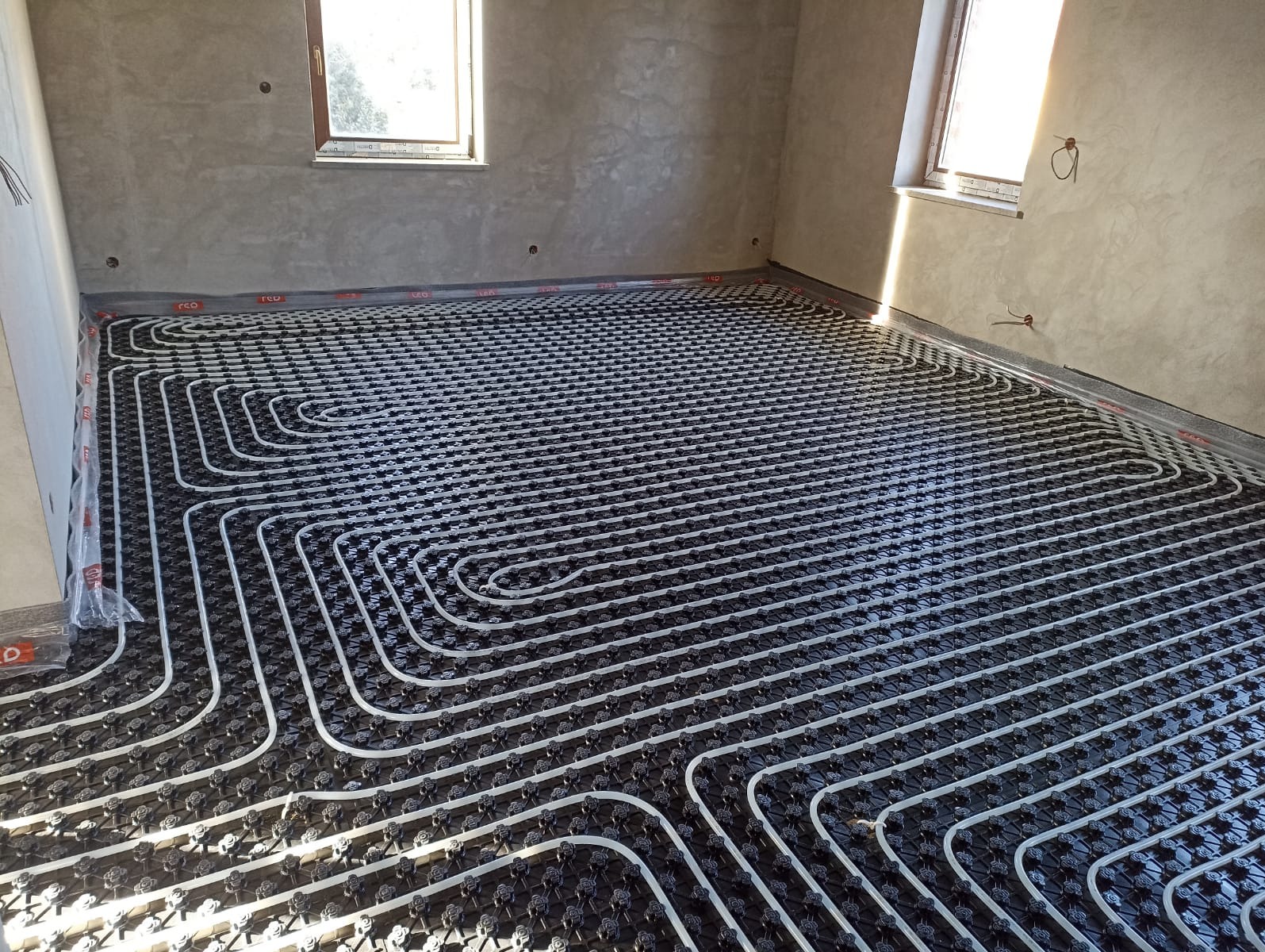 Białe rury ułożone w pętle na czarnej macie z wypustkami, przygotowane do wylewki pod ogrzewanie w surowym pomieszczeniu z oknami.