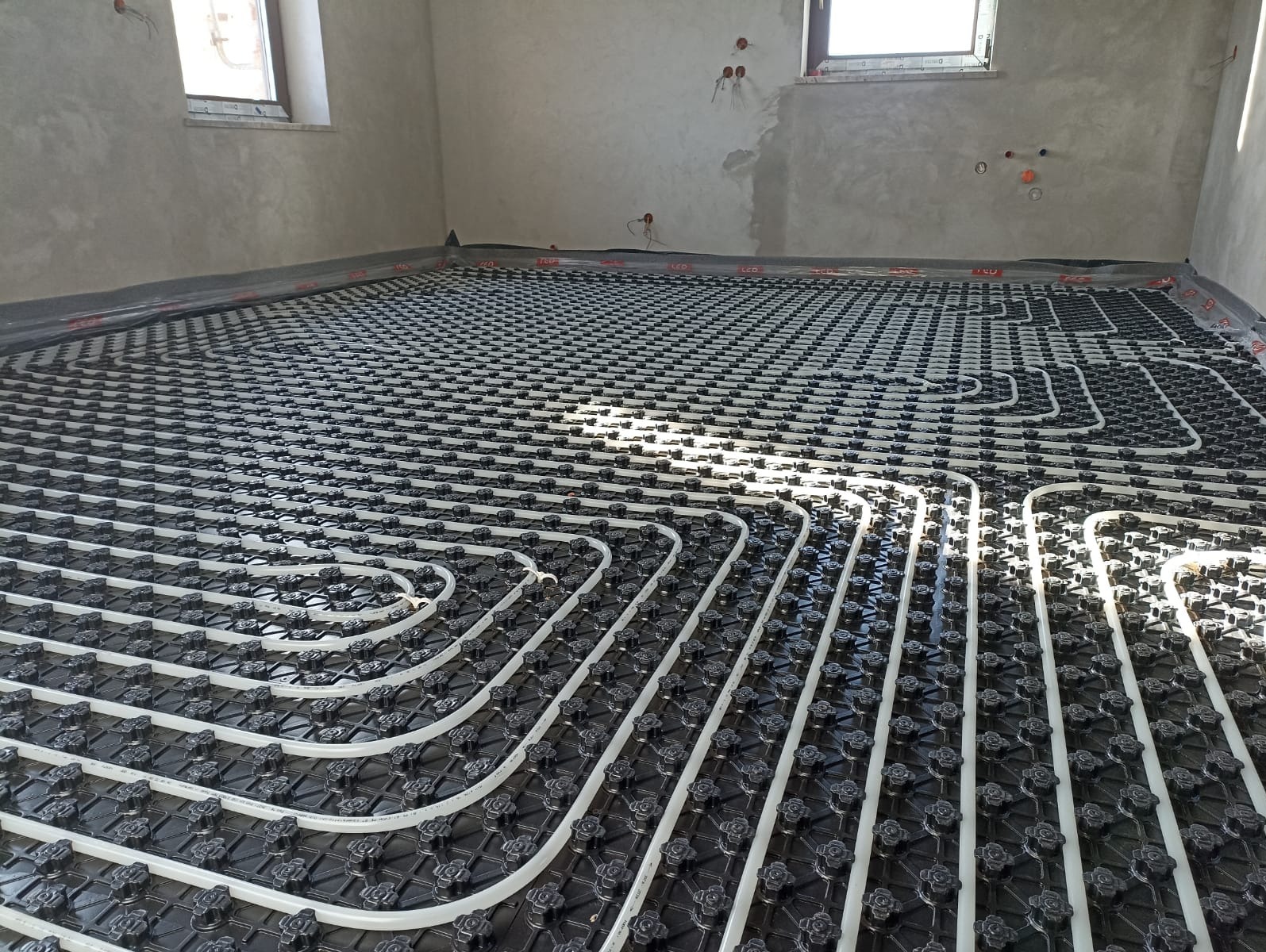 Białe rury ułożone w pętle na czarnej macie z mocowaniami, instalacja ogrzewania podłogowego w surowym pomieszczeniu z oknem i widocznymi przewodami elektrycznymi.