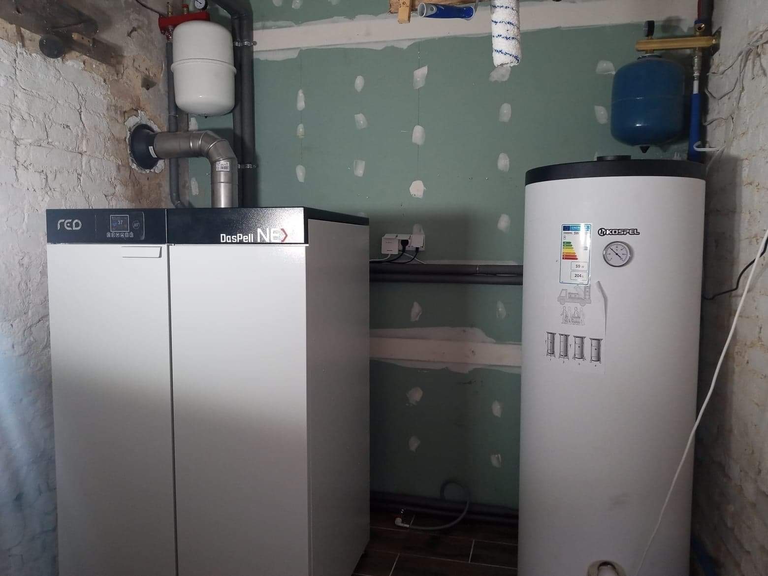 Nowoczesny piec gazowy Daspell Nex i bojler Kospel z niebieskim zbiornikiem wyrównawczym w surowym pomieszczeniu z częściowo wykończonymi ścianami.