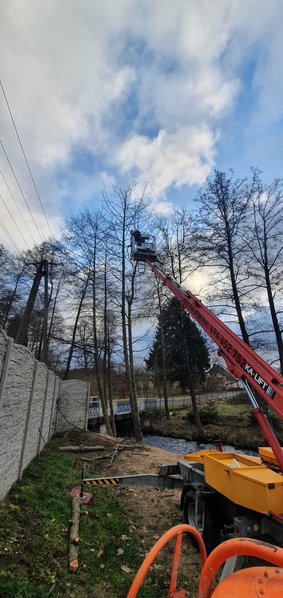 Pracownik na podnośniku koszowym przycina gałęzie drzew nad rzeką, widoczny betonowy płot i pocięte gałęzie na ziemi.