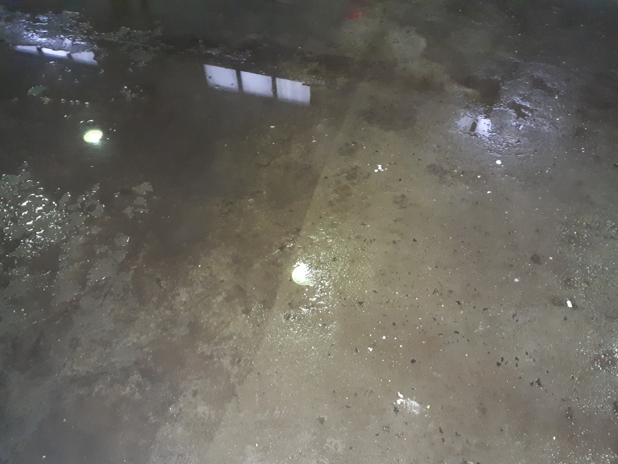 Mokra, brudna posadzka przemysłowa z refleksami świetlnymi i odbiciem okna, widoczne plamy i zabrudzenia.