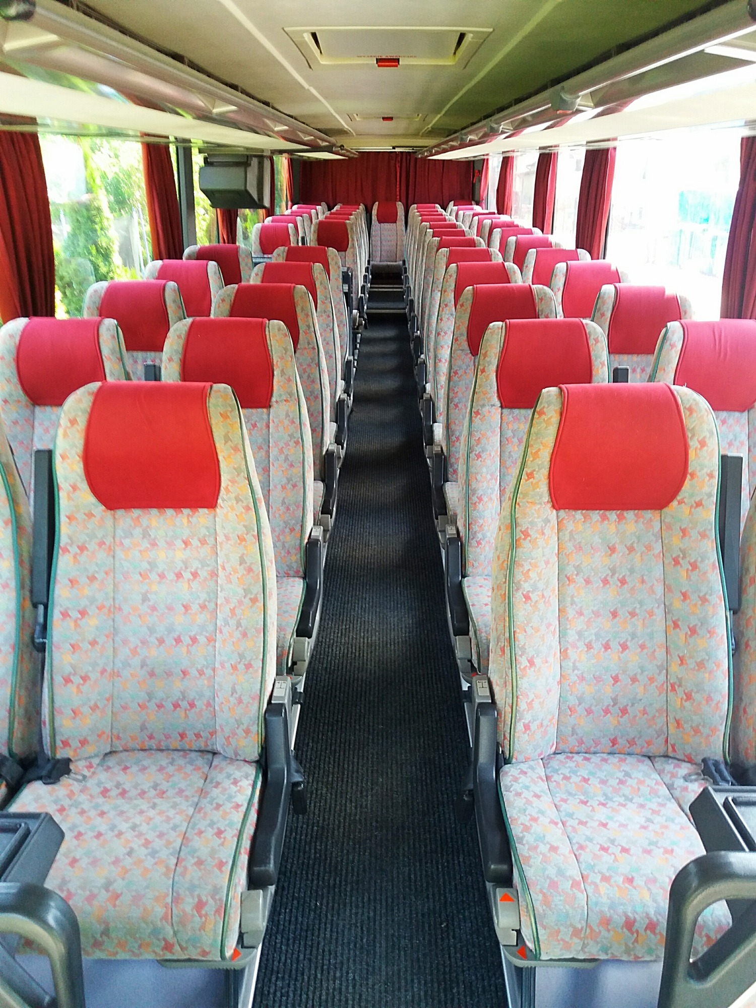 Wnętrze autokaru z rzędami foteli obitych wzorzystą tkaniną w odcieniach szarości, czerwone zagłówki, widok z przodu wzdłuż korytarza, zasłonięte okna.
