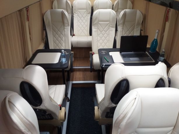 Wnętrze luksusowego busa z beżowymi skórzanymi fotelami, rozkładanymi stolikami, laptopem i butelką wody mineralnej na stoliku, sugerujące komfortową podróż.