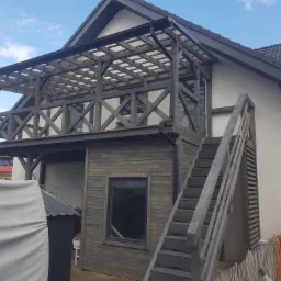 Dwukondygnacyjna konstrukcja z ciemnego drewna, zadaszona kratownicą, przylegająca do jasnego budynku z ciemnym dachem, widoczne schody zewnętrzne.