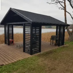 Czarna, drewniana altana z dachem krytym gontem bitumicznym, ustawiona na drewnianym podeście nad brzegiem jeziora, z widocznym grillem i czerwonym krzesłem wewnątrz.