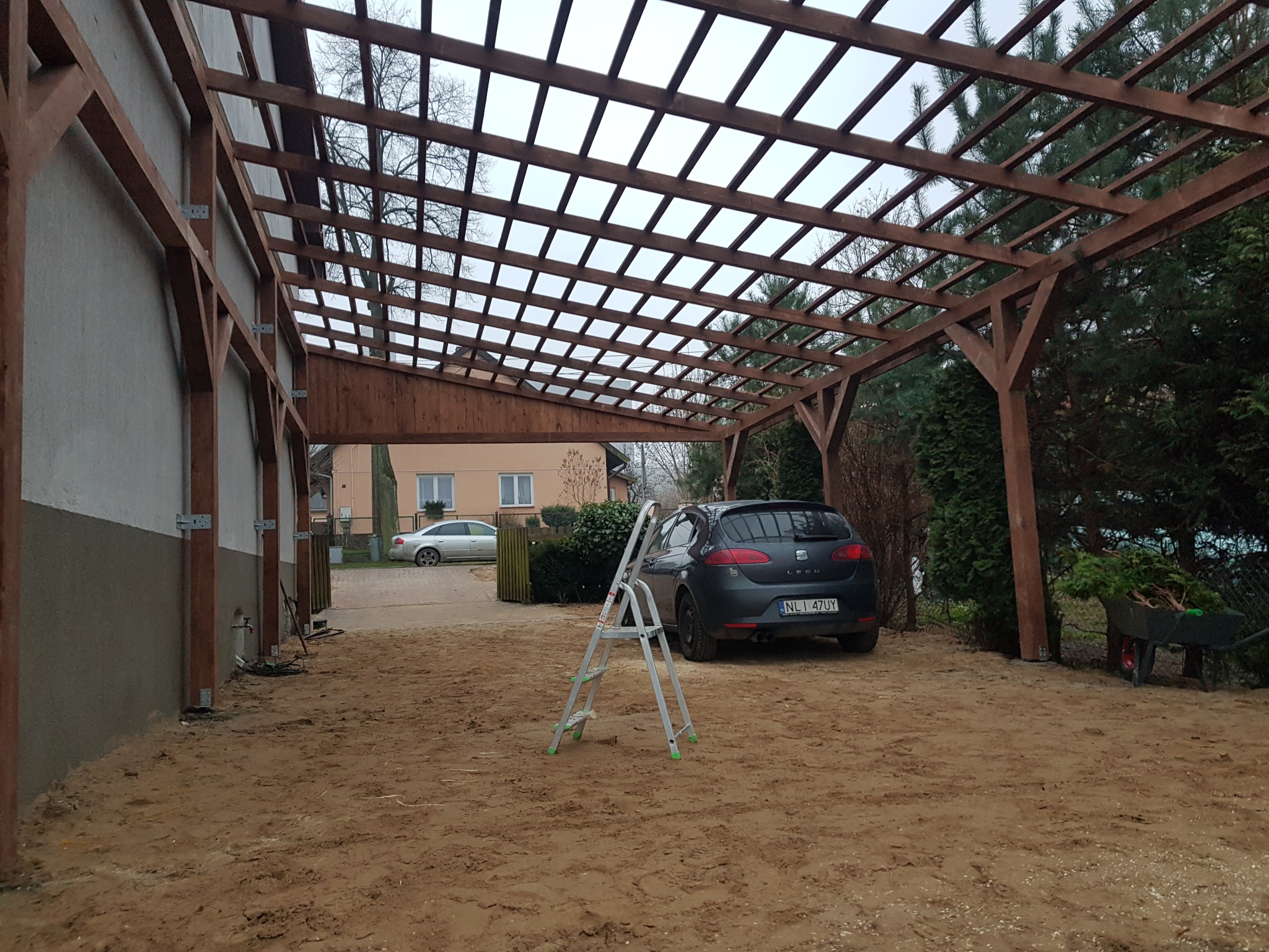 Drewniana konstrukcja z przezroczystym zadaszeniem w trakcie budowy, widoczna drabina, samochód i taczka w tle.