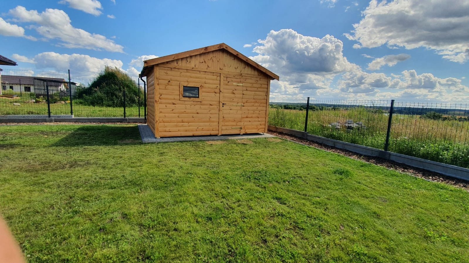 Drewniana altana ogrodowa z małym oknem, umieszczona na betonowej podstawie na trawniku, w tle błękitne niebo z białymi chmurami i ogrodzenie z siatki.