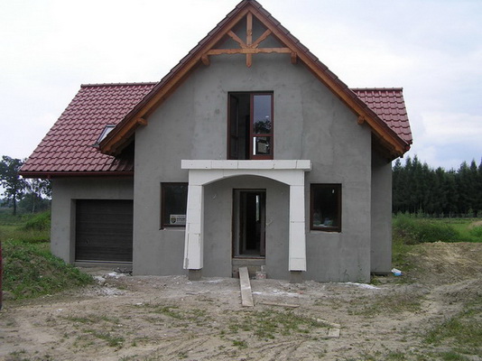 Surowy dom jednorodzinny w Dankowicach z nieotynkowaną elewacją, brązową dachówką i drewnianą konstrukcją więźby dachowej, widoczny biały, surowy portal wejściowy i brama garażowa.