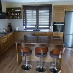 Jasna kuchnia z drewnianymi szafkami, blatem z ciemnego kamienia i trzema hokerami z brązowymi siedziskami przy wyspie.