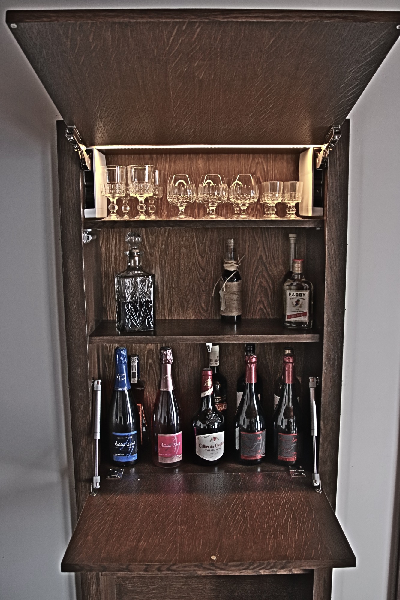 Drewniana szafka barowa z oświetleniem LED, otwierana do góry, wypełniona kryształowymi kieliszkami, karafką i butelkami alkoholu.