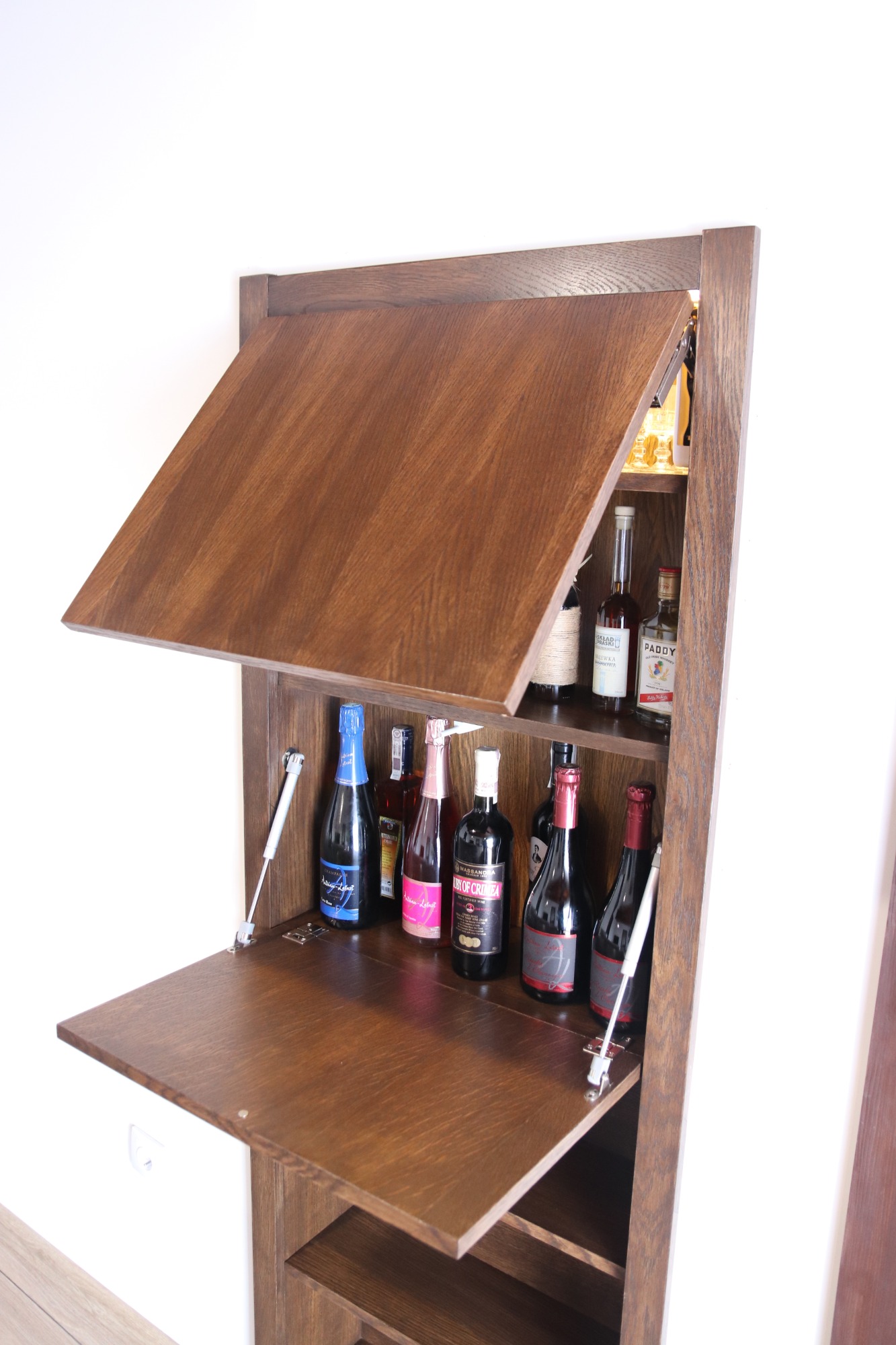 Drewniana szafka barowa na wymiar z otwieranym blatem i widocznymi butelkami alkoholu wewnątrz.