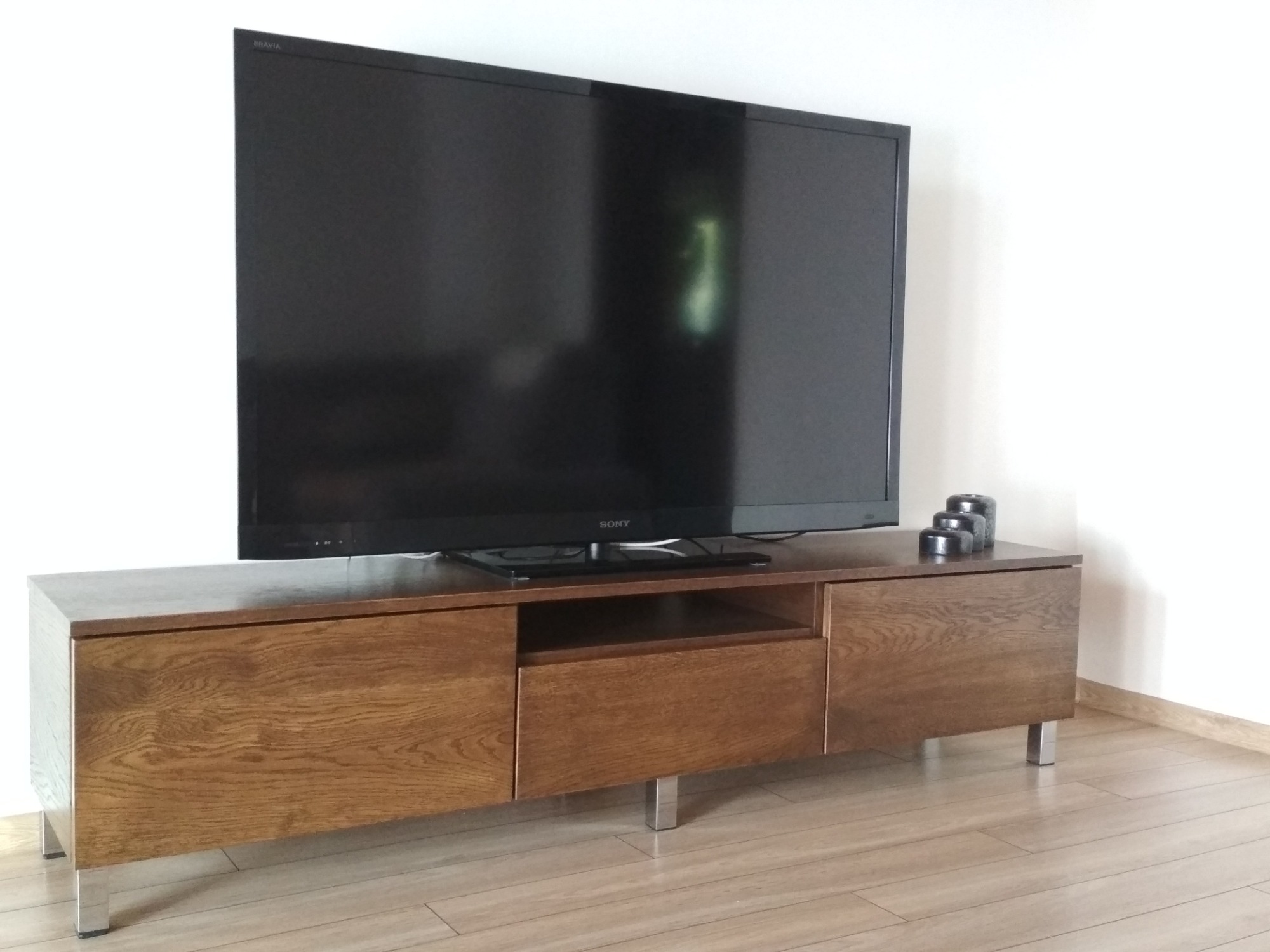 Drewniana szafka RTV z szufladami i otwartą półką, wsparta na metalowych nóżkach, z telewizorem Sony na blacie i trzema dekoracyjnymi elementami obok.