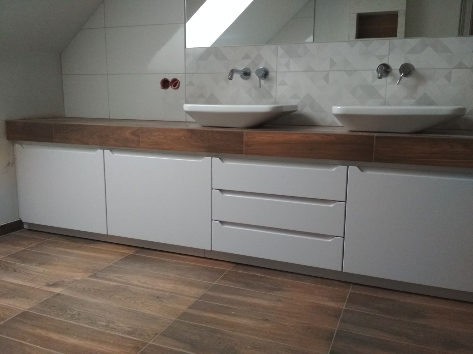 Łazienka na poddaszu z dwiema umywalkami nablatowymi, drewnianym blatem i białymi szafkami z szufladami, wykończona płytkami w geometryczny wzór.