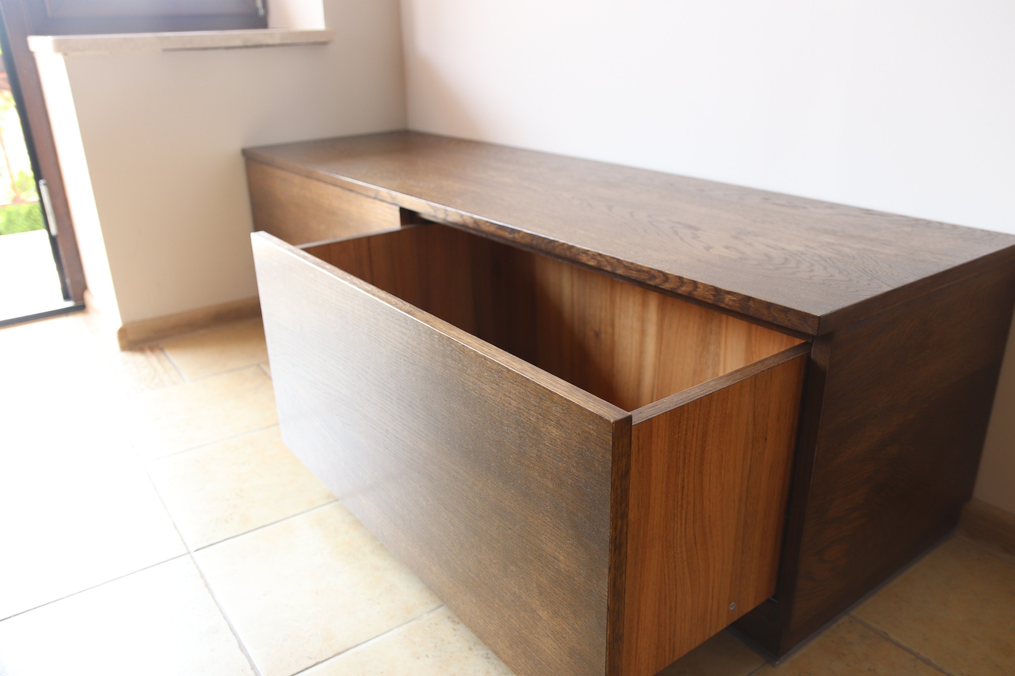 Drewniana komoda z wysuwaną szufladą, widoczne usłojenie drewna, jasne płytki podłogowe.