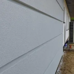 Świeżo otynkowana elewacja budynku jednorodzinnego z dekoracyjnymi poziomymi wgłębieniami, widok na narożnik, z zamontowaną jednostką zewnętrzną klimatyzacji i piaszczystym terenem w tle.