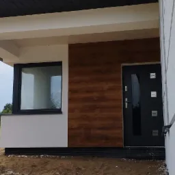 Fragment elewacji domu jednorodzinnego z drewnopodobną okładziną, białymi kolumnami werandy i czarnymi drzwiami wejściowymi, widok z zewnątrz na jeszcze niezagospodarowany teren.