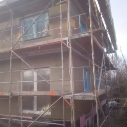 Dom w trakcie ocieplania, widoczne rusztowanie, okna zabezpieczone folią, ściany pokryte szarym tynkiem, na dole różowe płyty izolacyjne.