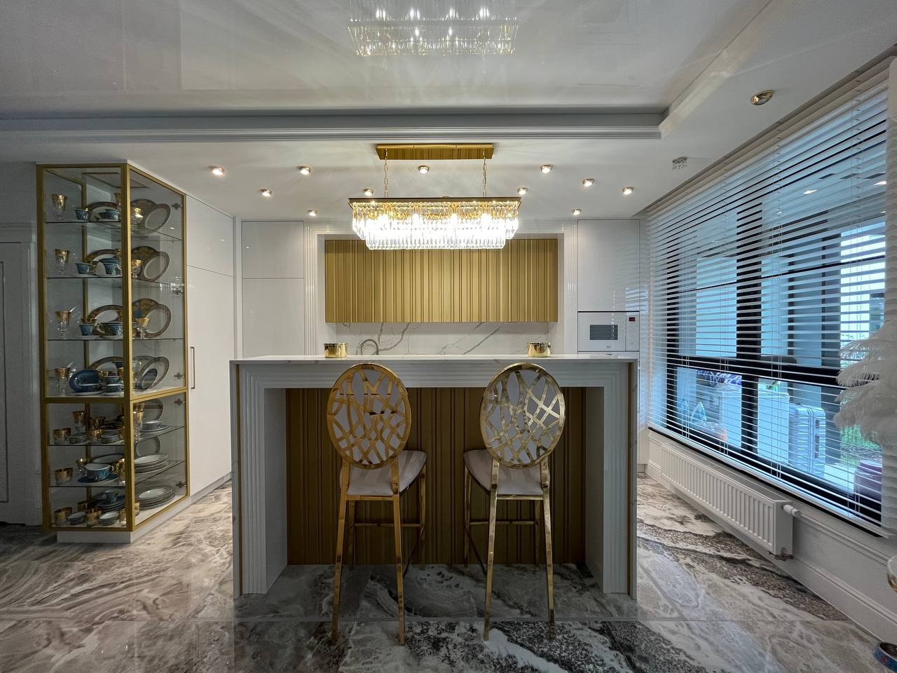 Elegancka kuchnia z białymi szafkami, złotymi akcentami i marmurową podłogą; wyspa kuchenna z dwoma złotymi krzesłami barowymi i oświetleniem wiszącym.