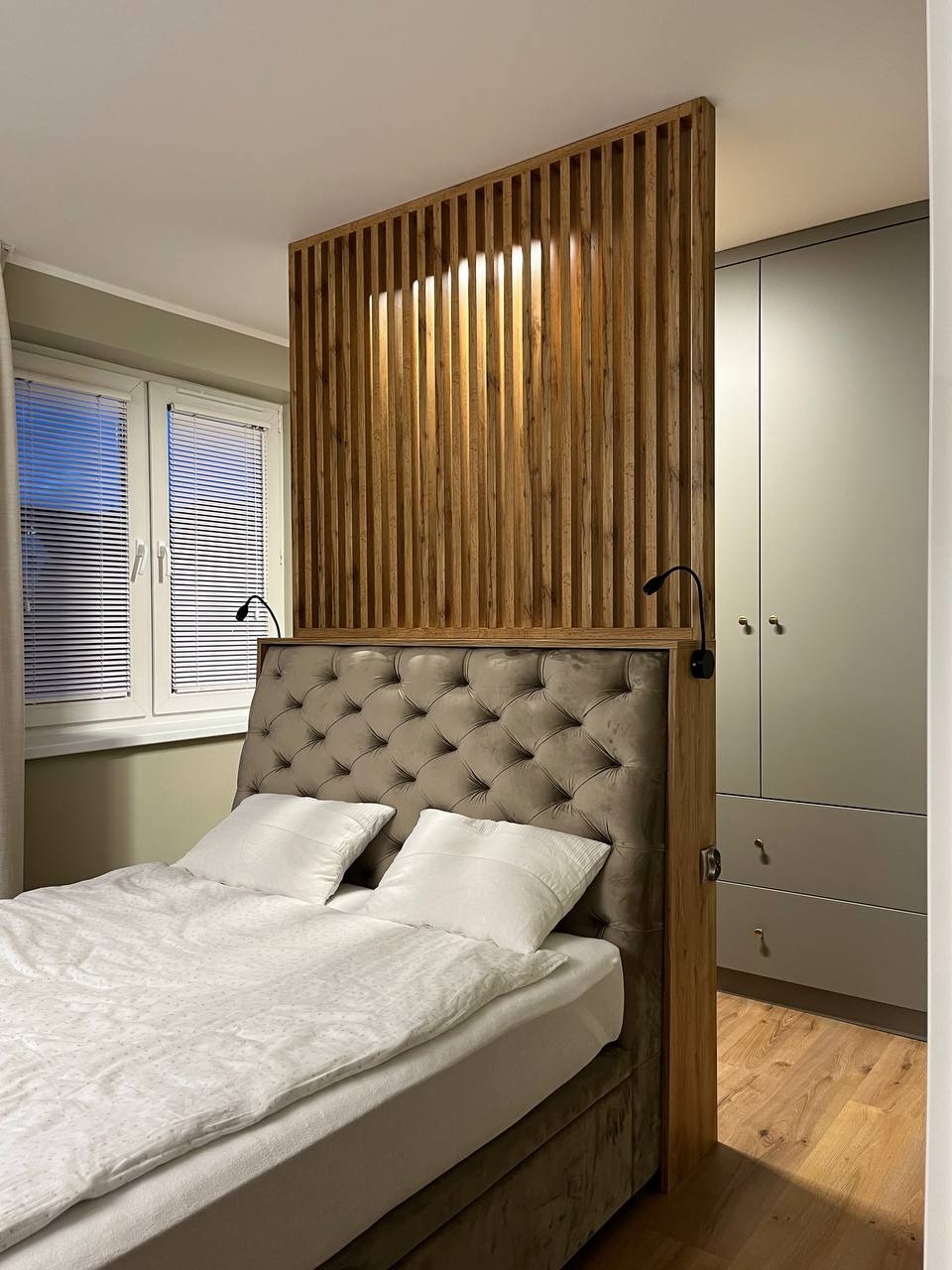 Nowoczesna sypialnia z łóżkiem z pikowanym zagłówkiem i drewnianą ażurową ścianką działową z oświetleniem, obok szafa w odcieniach szarości.