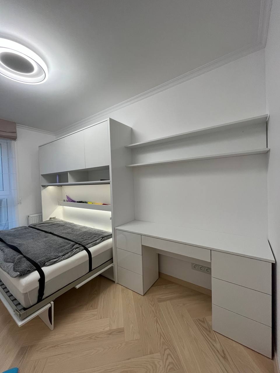 Biała zabudowa meblowa z rozkładanym łóżkiem, biurkiem i półkami, widok na łóżko z szarą narzutą, parkiet w jodełkę na podłodze.