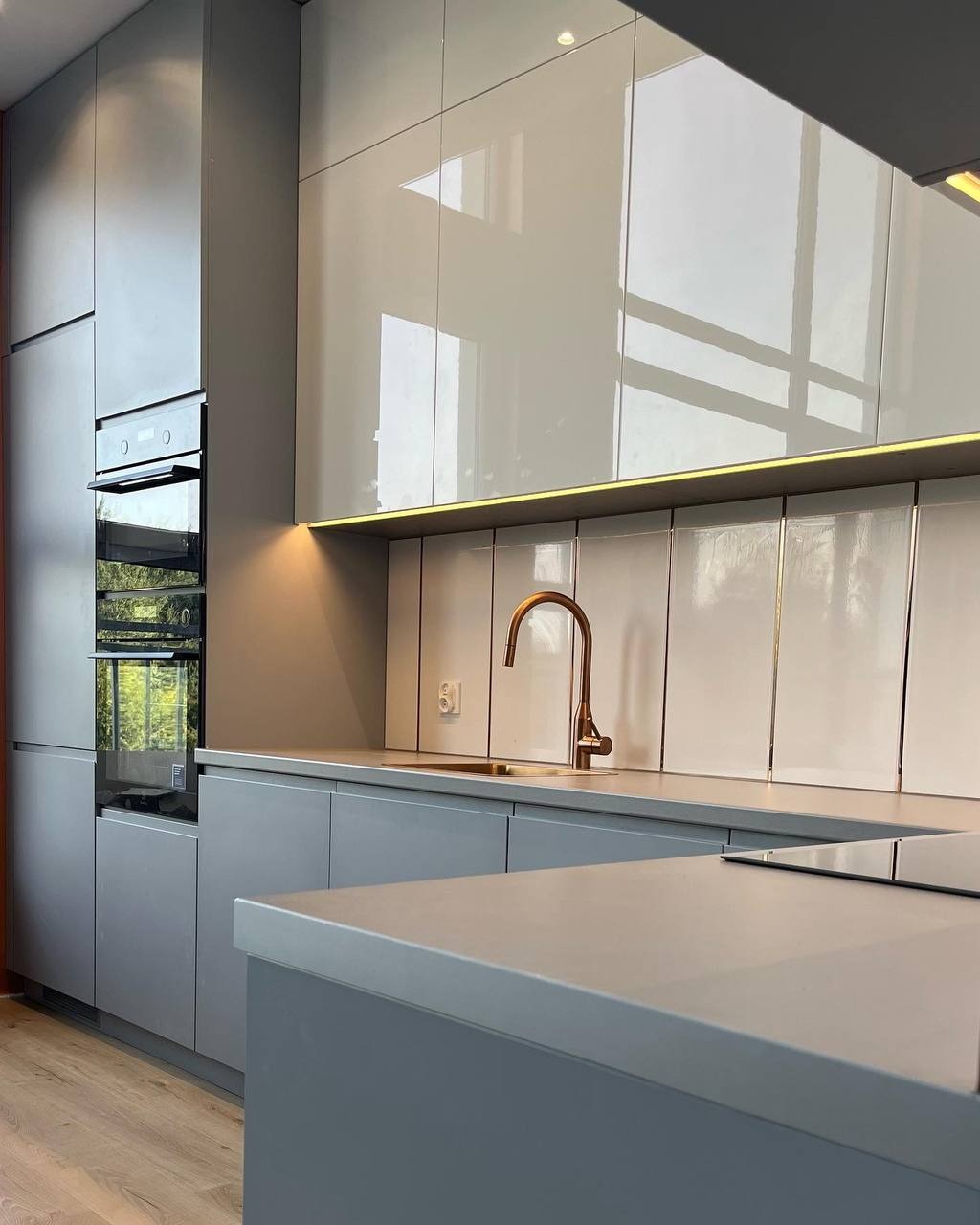 Nowoczesna kuchnia w odcieniach szarości z błyszczącymi frontami szafek, złotą armaturą i subtelnym oświetleniem LED pod szafkami wiszącymi.