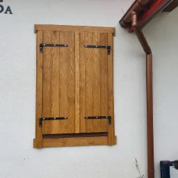 Drewniane okiennice zewnętrzne z czarnymi zawiasami na elewacji budynku, widoczny numer domu 45A i rynna spustowa.