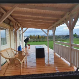 Zadaszony drewniany taras przy domku letniskowym z drewnianą podłogą i balustradą, na którym stoi rattanowy fotel, butla gazowa i czarny stolik; kobieta w oddali zamiata taras.