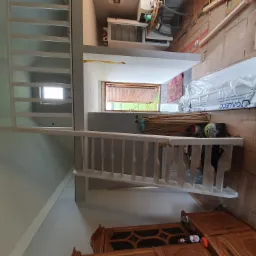 Wnętrze w trakcie remontu z widoczną białą antresolą z drewnianymi schodami, częściowo zamontowaną, z widocznymi materiałami budowlanymi i narzędziami w tle.