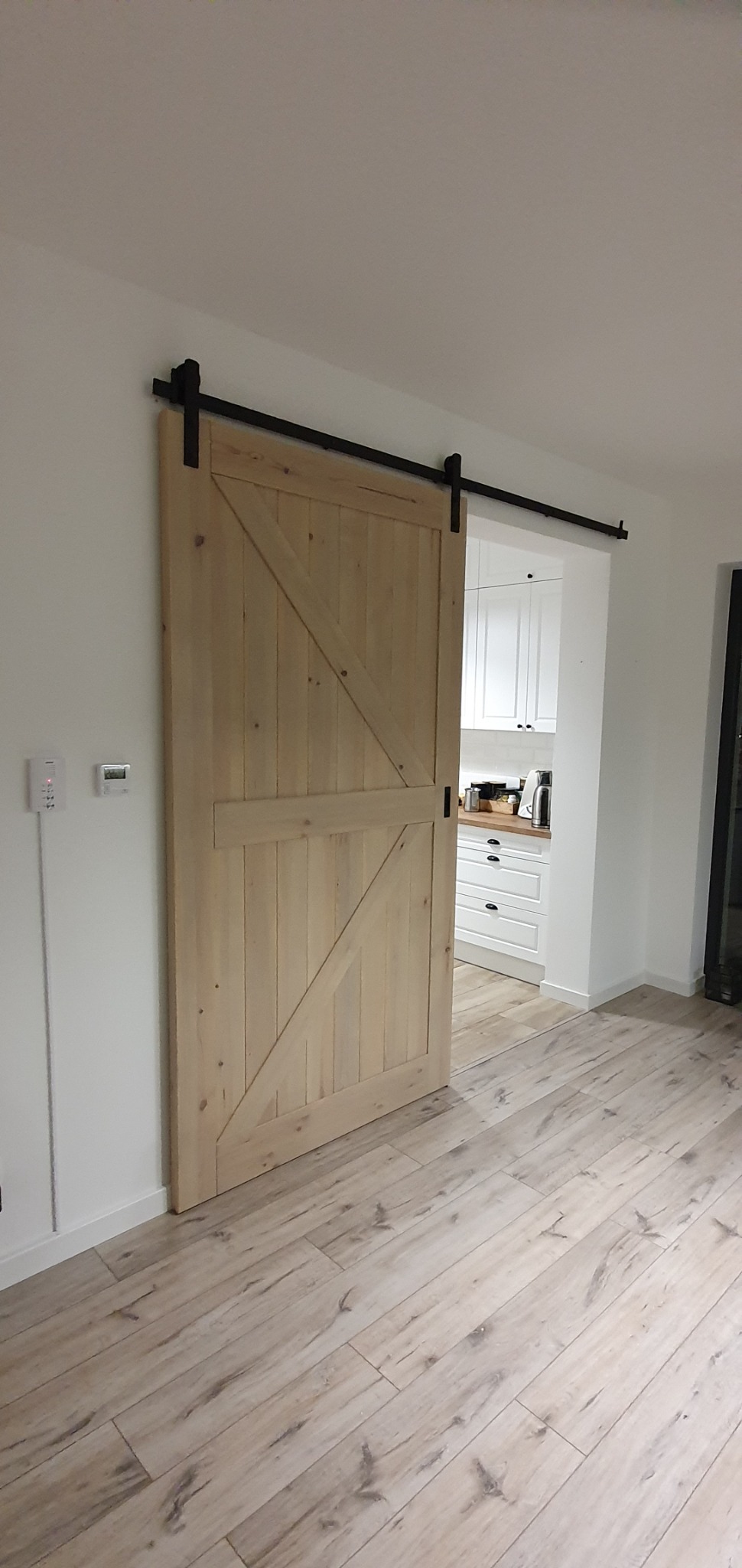 Drewniane drzwi przesuwne w stylu rustykalnym, z widocznym systemem przesuwnym na czarnej szynie, częściowo odsłaniające wnętrze kuchni z białymi meblami.