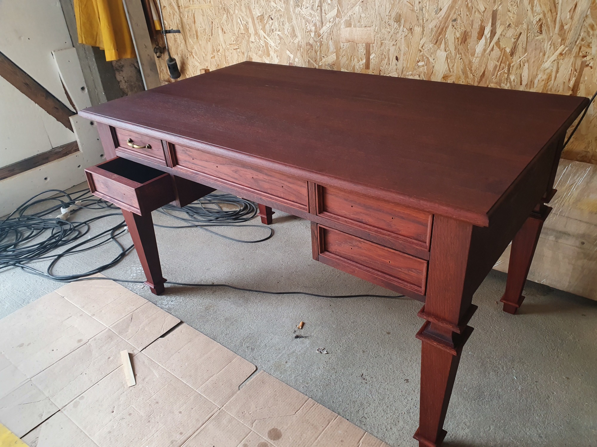 Biurko z drewna w odcieniu mahoniu z wysuniętą szufladą, widoczne kable i karton na podłodze w warsztacie.