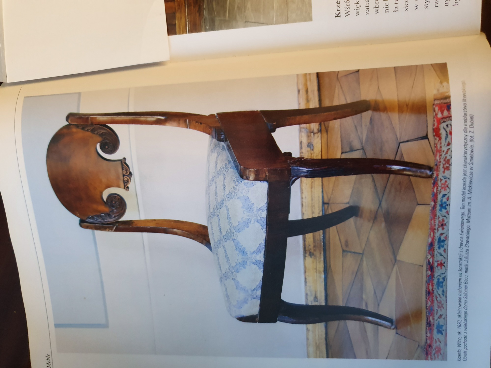 Zdjęcie krzesła z epoki, mahoniowe okleinowanie, widoczny wzór tapicerki siedziska, na tle parkietu i fragmentu tekstu opisującego obiekt muzealny.