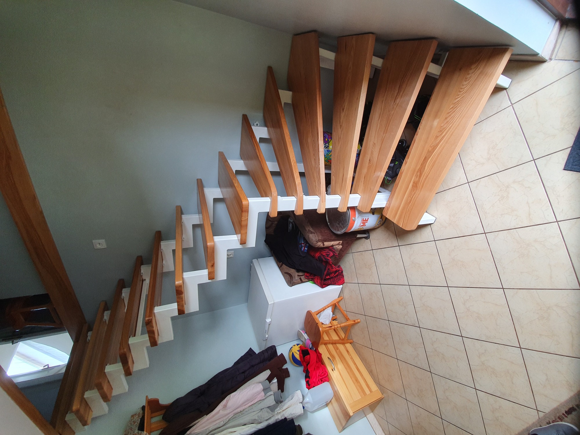 Nowoczesne schody drewniane z białymi wspornikami, widok z góry na tle beżowej podłogi, z widocznym bałaganem pod schodami.