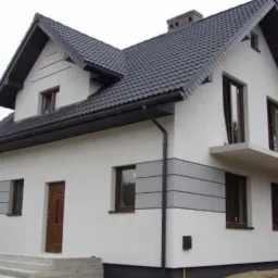 Dwukondygnacyjny dom jednorodzinny z szarą dachówką, białymi ścianami i szarymi akcentami dekoracyjnymi na elewacji, widoczny z zewnątrz, po remoncie.