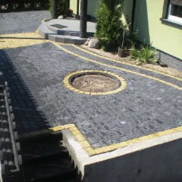 Ułożona nawierzchnia z szarej kostki brukowej z żółtymi obrzeżami, widoczny okrągły obszar przygotowany pod ognisko, schody i fragment ogrodzenia.