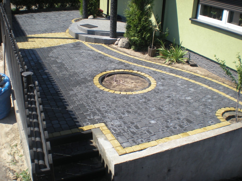 Ułożona nawierzchnia z szarej kostki brukowej z żółtymi obrzeżami, widoczny okrągły obszar przygotowany pod ognisko, schody i fragment ogrodzenia.