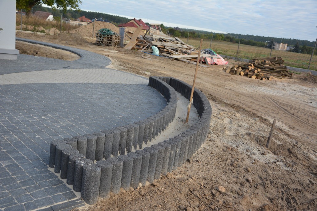 Ułożona szara kostka brukowa z okrągłym obrzeżem z betonowych palisad, widoczne przygotowane podłoże i składowane materiały budowlane w tle.