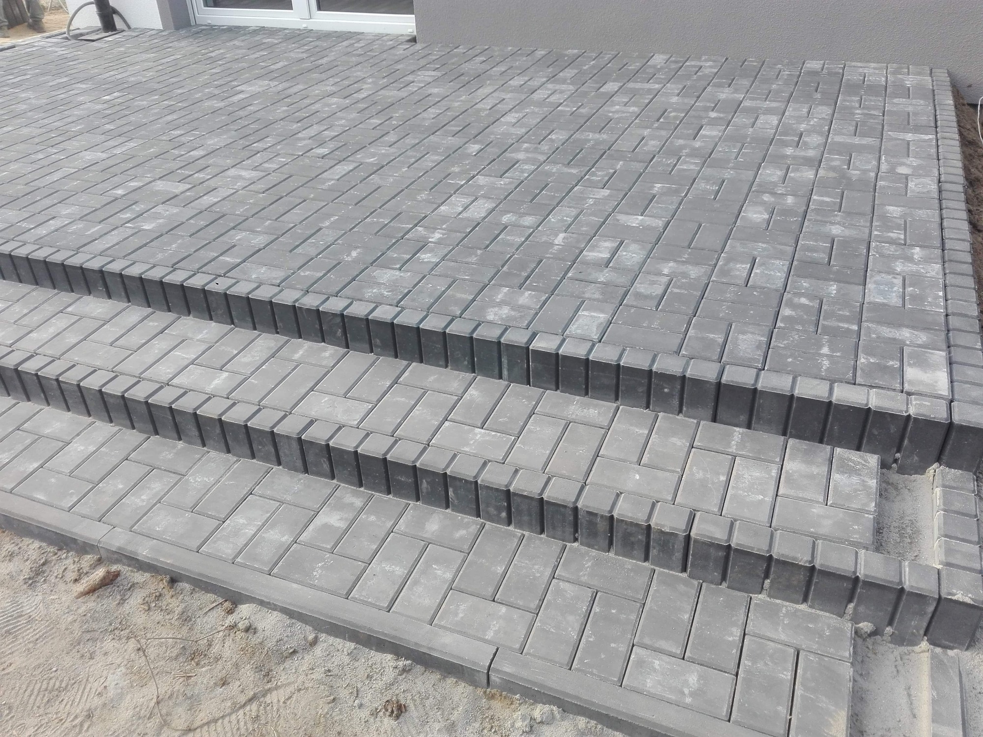 Schody wejściowe wyłożone szarą kostką brukową w jodełkę, z obrzeżami z kostki pionowej, widoczne niedociągnięcia w postaci piasku na krawędziach.