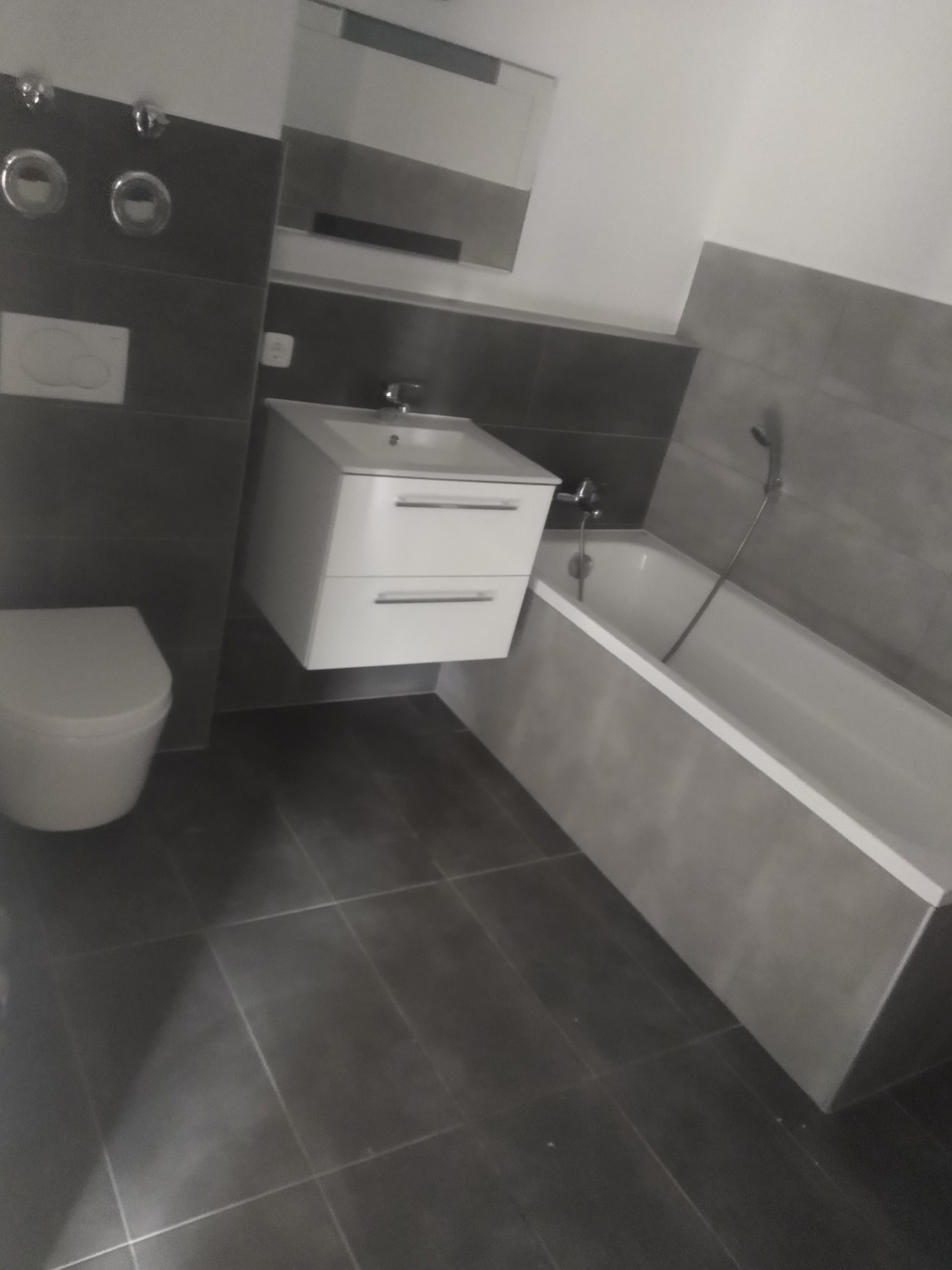 Nowoczesna łazienka z szarymi płytkami, podwieszaną szafką z umywalką i białą wanną.