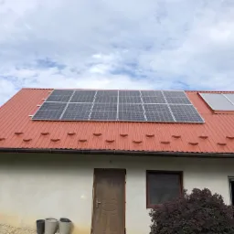 Dom jednorodzinny z panelami słonecznymi na czerwonym dachu, widoczne trzy donice przed budynkiem i krzew z bordowymi liśćmi.