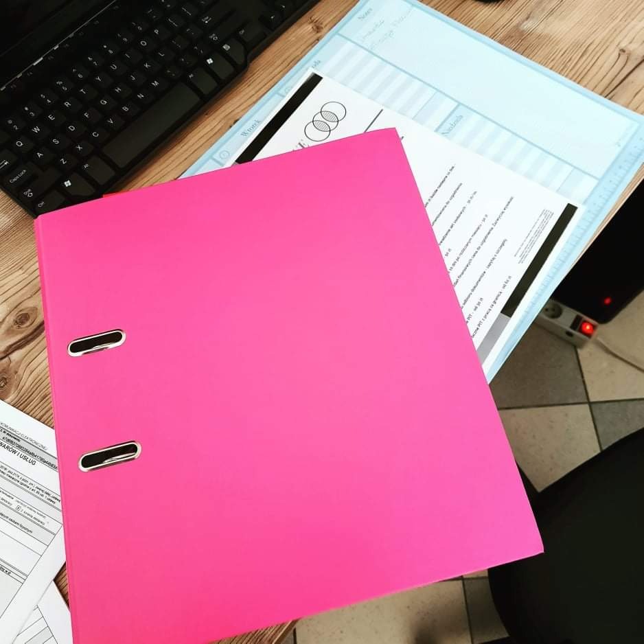 Różowy segregator na drewnianym biurku obok klawiatury, dokumentów i podkładki. Segregator zawiera metalowe okucia. Dokumenty częściowo widoczne, jeden z nich z nagłówkiem 'Wtorek'.