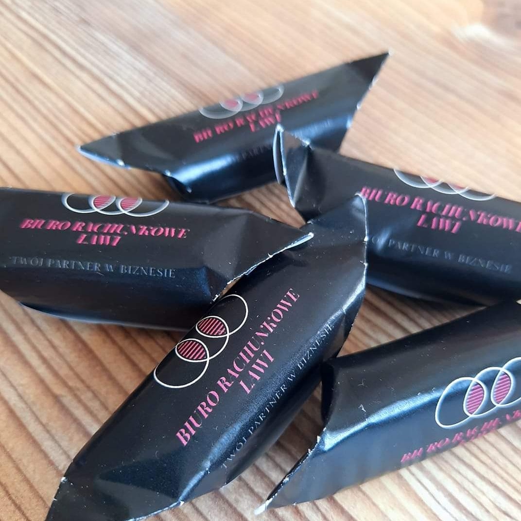 Czarne, błyszczące cukierki reklamowe z nadrukiem nazwy i logo firmy na drewnianym tle.