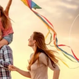 Uśmiechnięta rodzina z dzieckiem puszczającym kolorowego latawca na tle słonecznego nieba, logo firmy w prawym górnym rogu.