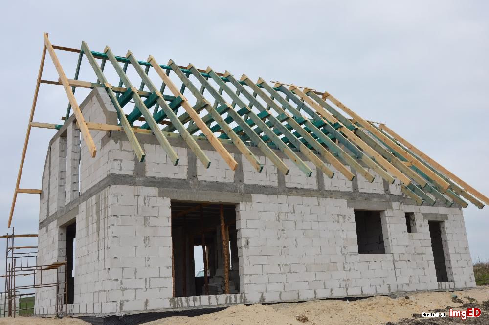 Surowy stan domu murowanego z betonu komórkowego, z widoczną konstrukcją drewnianą dachu w trakcie montażu, bez okien i drzwi, na piaszczystym podłożu.