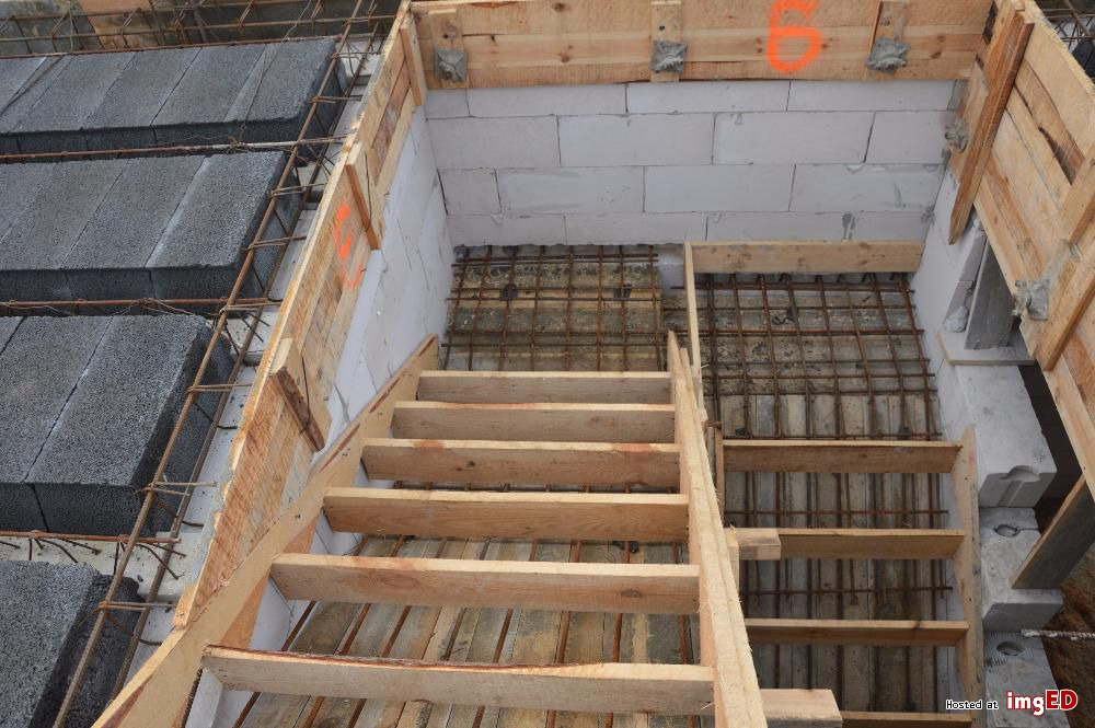 Wnętrze domu w trakcie budowy: widoczne bloczki z betonu komórkowego, drewniane szalunki, zbrojenie i tymczasowe schody drewniane.